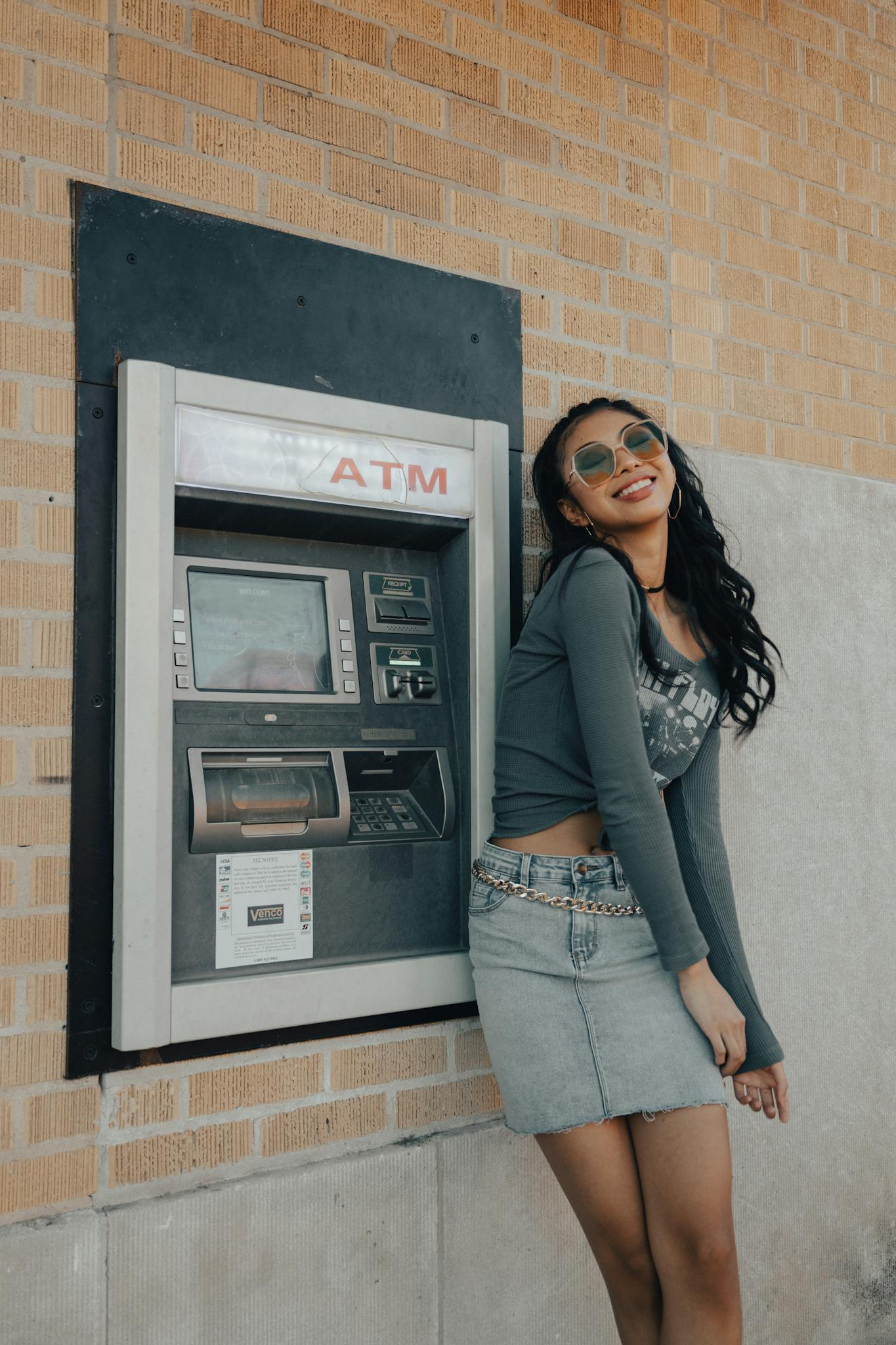A young woman in trendy fashion posing happily by an outdoor ATM machine.