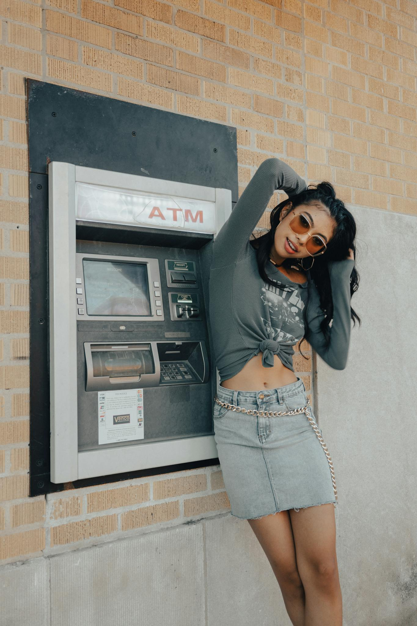 Fashionable woman posing by an ATM on a sunny day wearing a denim skirt and sunglasses.