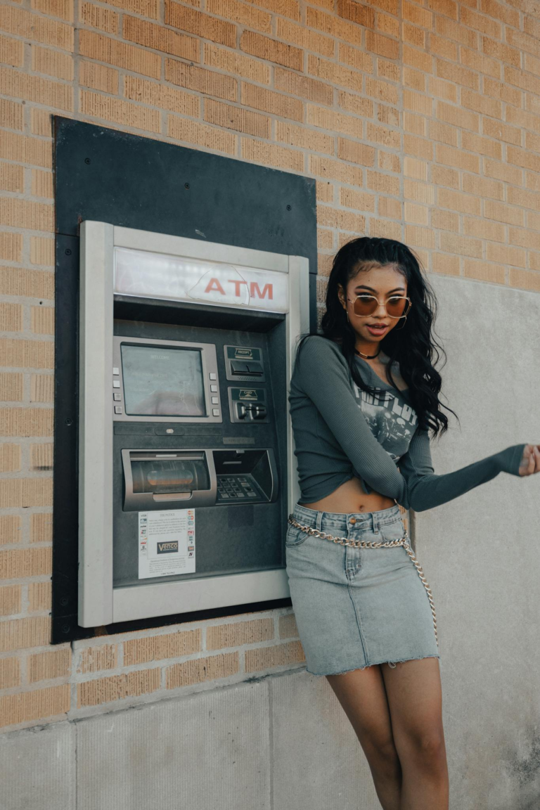 Stylish young woman standing beside an outdoor ATM in a city setting.